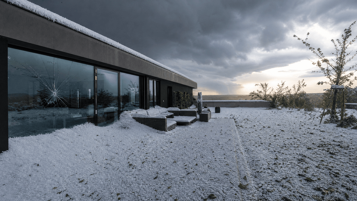 Glasfront eines modernen Hauses mit zersprungener Scheibe, Terrasse und Garten voller weißer Hagelkörner
